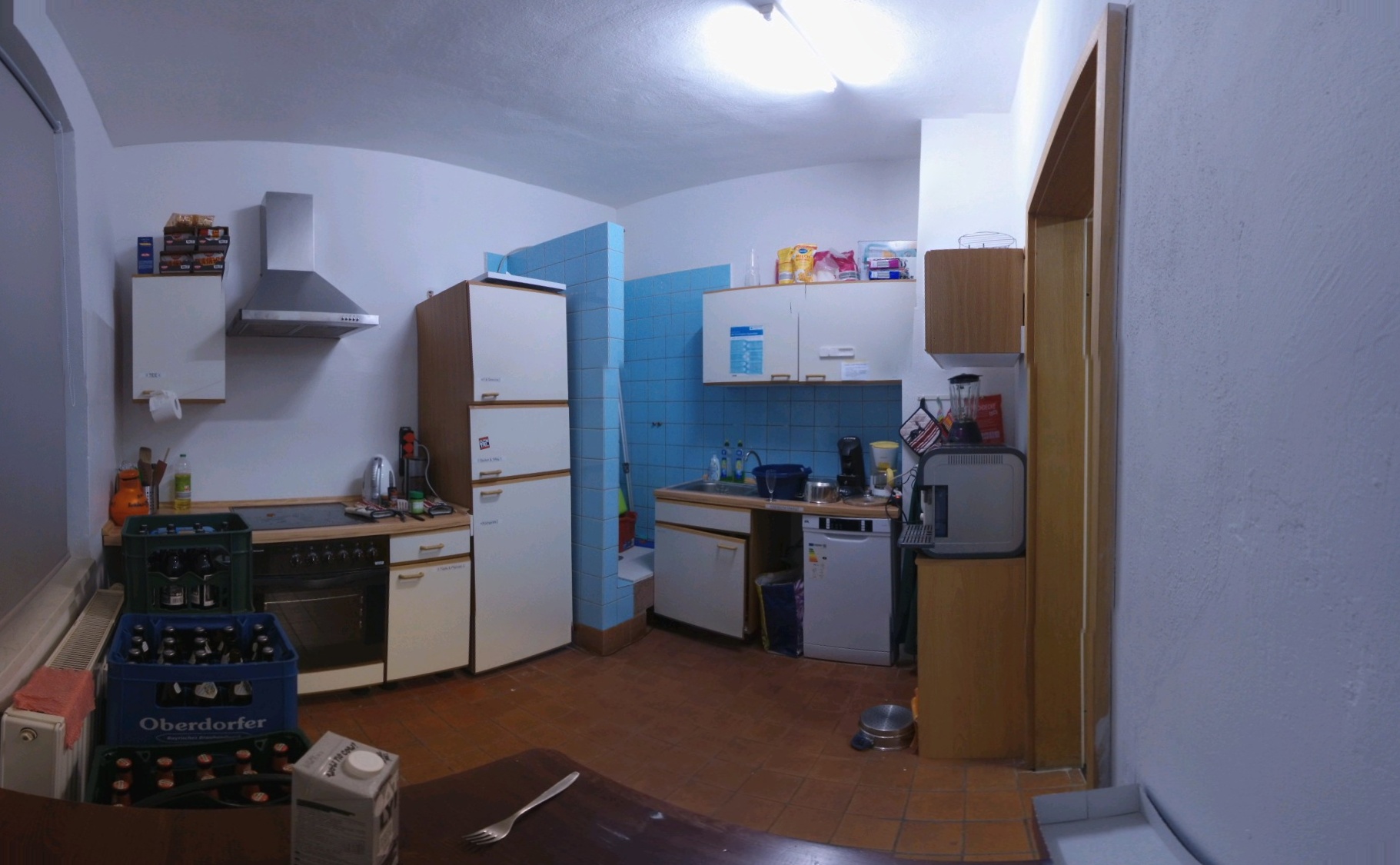 Eine unaufgeräumte Küche, welche einen blau gefließten Bereich enthält, welcher wie eine Dusche aussieht. Mitten in der Küche! Alles zum Kochen ist vorhanden, ein Nudelabgießtopf liegt auf dem Boden, eine Gabel und Milchpackung vor der Kamera und eine Toilettenpapierrolle wurde als Aufwischtücherersatz installiert